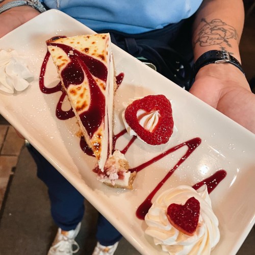 A slice of cheesecake topped with raspberry sauce, garnished with strawberries and whipped cream, served on a plate.