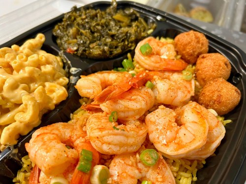 A close-up of a meal featuring shrimp, rice, macaroni and cheese, greens, and fried balls in a black takeout container.