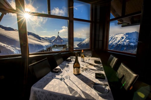 A beautifully set dining table with wine, overlooking snow-covered mountains and a bright sunlit sky through large windows.