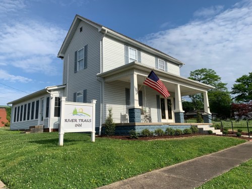 A white two-story inn with an American flag, surrounded by green grass and a sign that reads "River Trails Inn."