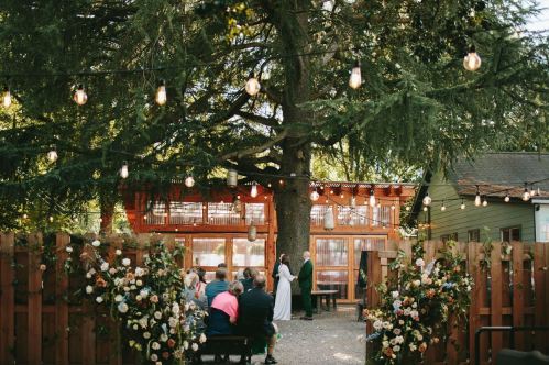 A wedding ceremony under a large tree, with guests seated and floral decorations, string lights overhead.