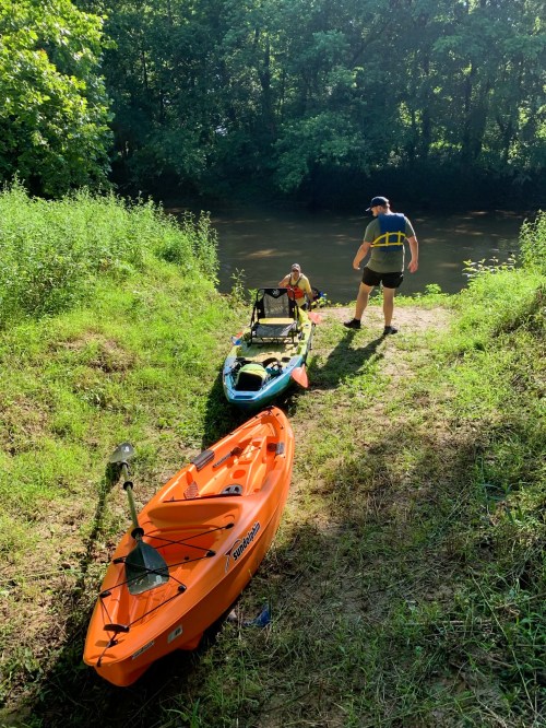 Two kayaks on a riverbank, with one person preparing to launch and another seated in a kayak. Lush greenery surrounds them.
