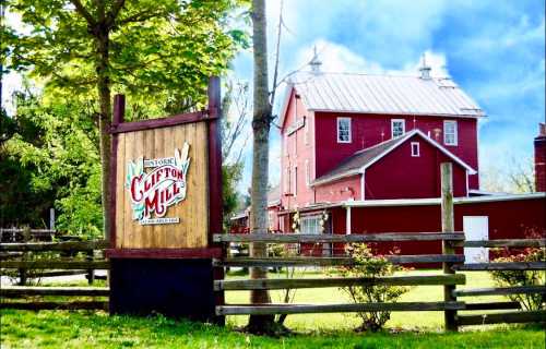 A red historic mill building with a wooden sign reading "Clifton Mill," surrounded by greenery and a blue sky.