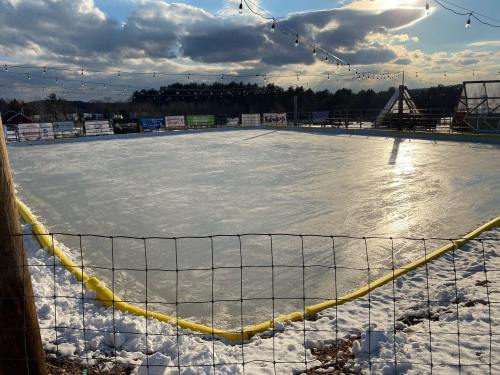 An empty outdoor ice rink surrounded by snow, with a cloudy sky and string lights in the background.