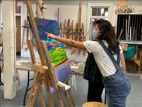 Two people in an art studio, one pointing at a colorful painting on an easel while wearing masks.