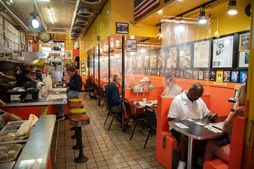 A vibrant diner interior with colorful walls, booths, and patrons enjoying meals, separated by clear plastic barriers.