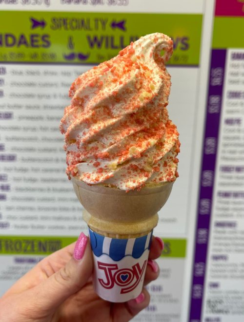 A hand holding a soft-serve ice cream cone topped with red sprinkles against a colorful menu background.