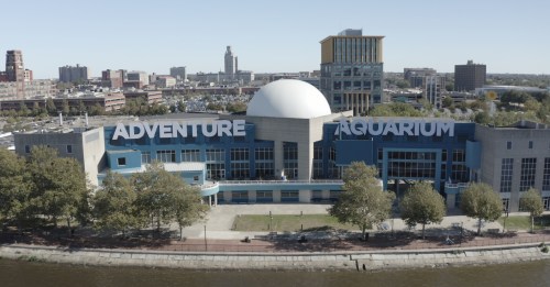 Aerial view of Adventure Aquarium, featuring a large dome and blue exterior, surrounded by trees and a river.
