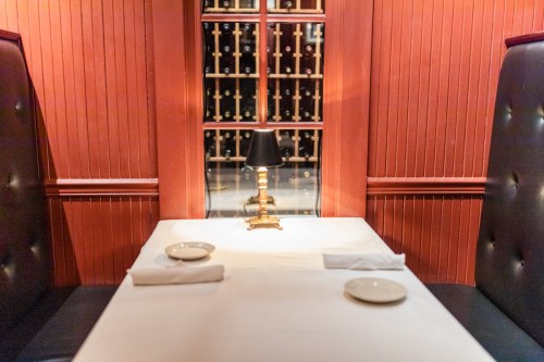 A cozy restaurant table set for two, featuring a small lamp and wine bottles in the background.