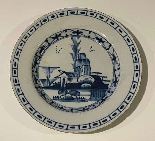A decorative blue and white plate featuring a landscape scene with trees, a house, and stylized hills.
