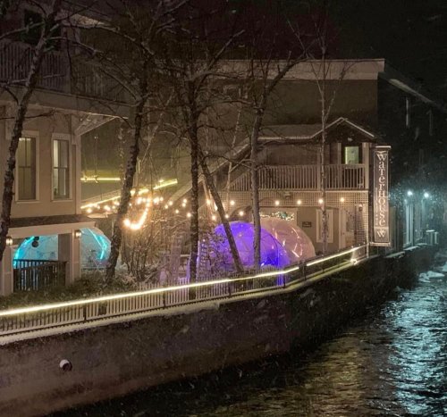 A cozy riverside restaurant with illuminated igloo-like structures and festive lights, surrounded by snow.