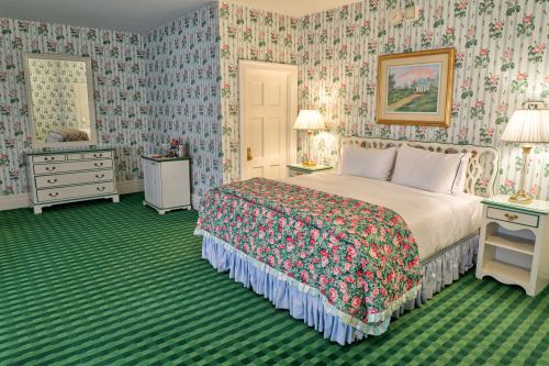 A cozy bedroom with floral wallpaper, a patterned bedspread, and green striped carpet, featuring a dresser and mirror.