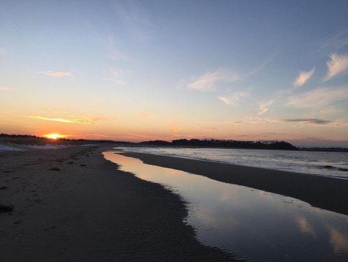 A serene beach at sunset, with gentle waves and a reflective pool of water along the shore.