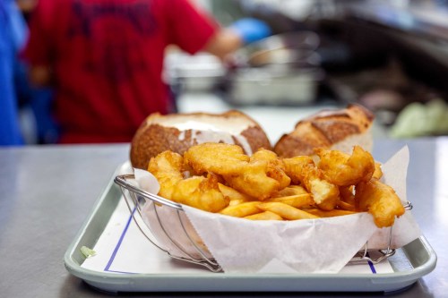 A tray with a basket of golden fried fish and fries, alongside two bread rolls in the background.