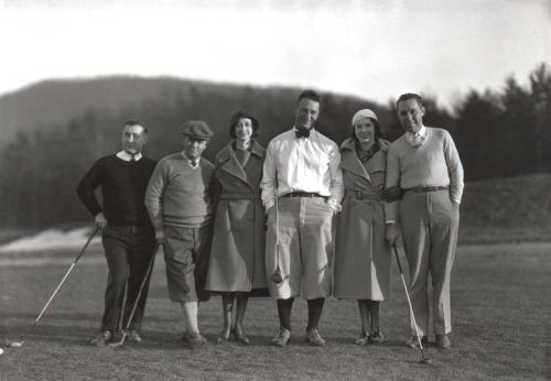 A group of six people in vintage golf attire pose together on a golf course, with hills in the background.