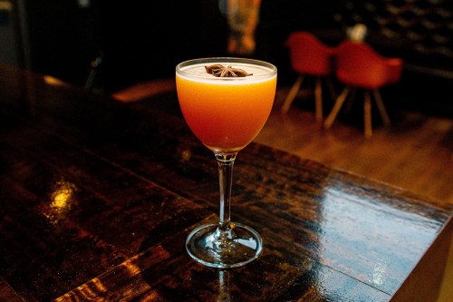 A cocktail in a stemmed glass, featuring a vibrant orange hue and garnished with star anise, on a polished wooden bar.