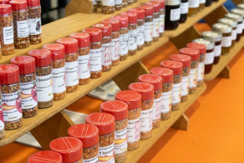 A display of various spice jars on a wooden shelf, each labeled with names and prices.