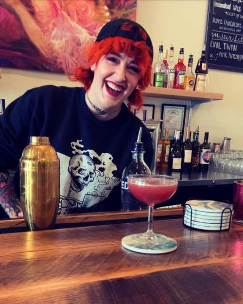 A bartender with bright orange hair smiles behind the bar, showcasing a cocktail and various bottles in the background.