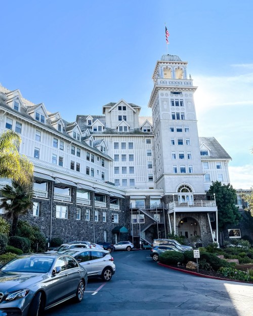 Historic hotel with a tall tower, surrounded by palm trees and parked cars, under a clear blue sky.