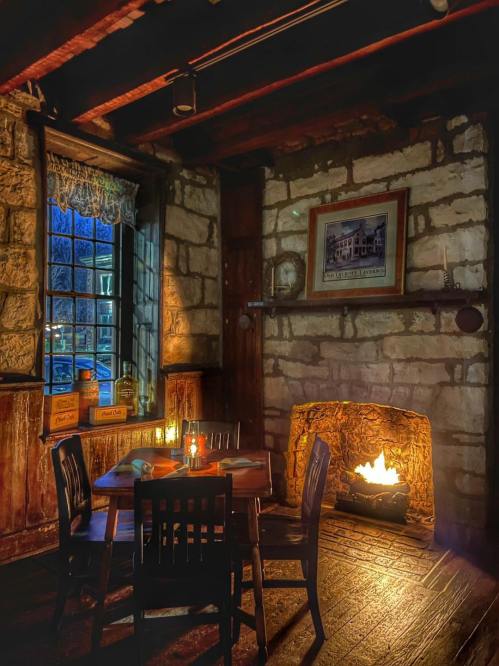 Cozy interior of a rustic room with a wooden table, chairs, and a glowing fireplace beside a window.