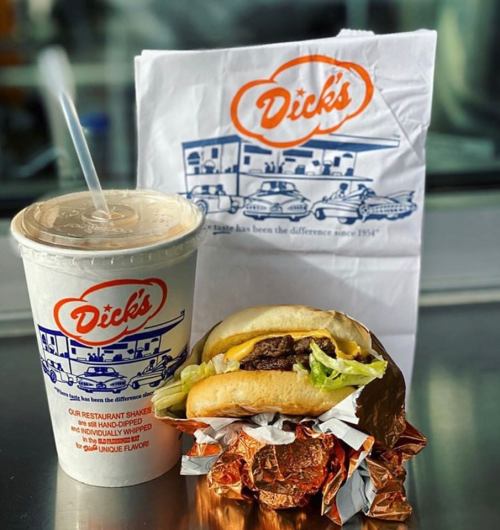 A burger wrapped in foil next to a cup of shake, with a branded paper bag in the background.
