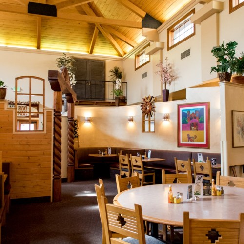 Cozy restaurant interior with wooden beams, round tables, and warm lighting, featuring plants and decorative artwork.