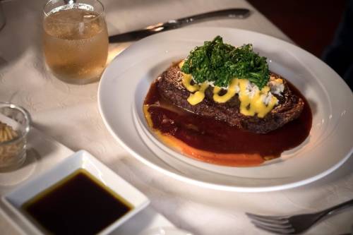 A plate of meatloaf topped with greens and cheese, served with a side of sauce and a drink in a glass.