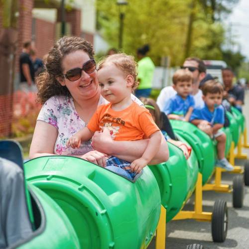 A woman smiles while holding a child on a green train ride, surrounded by other children enjoying the ride.
