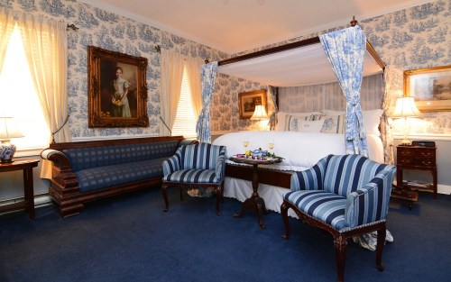 A cozy bedroom featuring a four-poster bed, patterned wallpaper, and elegant furniture, with soft lighting.