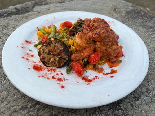 A plate of chicken in a rich sauce, served with roasted vegetables, cherry tomatoes, and a grilled lemon slice.