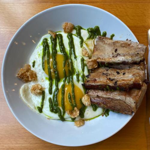 A bowl with two sunny-side-up eggs, green sauce drizzled on top, and slices of grilled meat beside crispy pork cracklings.
