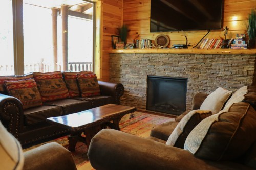 Cozy living room with a stone fireplace, wooden walls, and comfortable seating, featuring a coffee table and a TV.