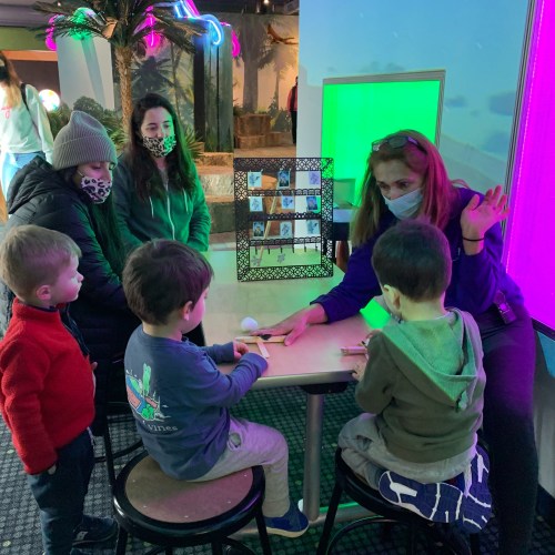 A group of children and adults gather around a table, engaging in an activity with colorful lights in the background.