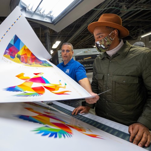 A man in a hat examines colorful prints with a printer in a workshop, both focused on the artwork.