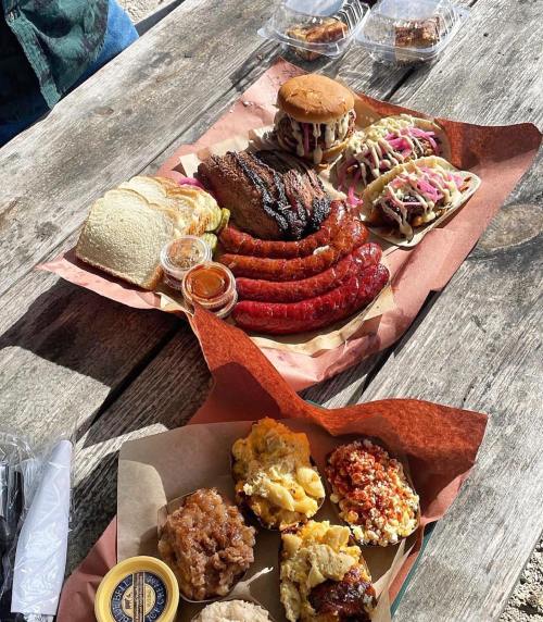 A spread of barbecue featuring sausages, brisket, a burger, and various sides on paper-lined trays.