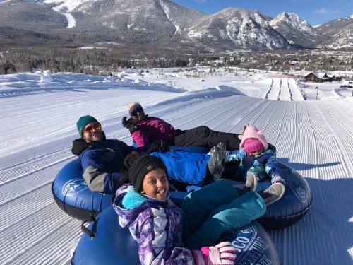A group of four people relax on snow tubes, surrounded by snowy mountains and a clear blue sky.