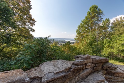 Scenic view from a rocky ledge, surrounded by trees, overlooking a valley and distant hills under a clear blue sky.