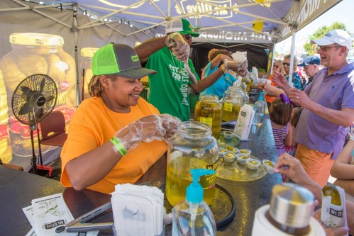 A vendor serves pickles at a festival booth, with customers eagerly reaching for samples in a lively outdoor setting.