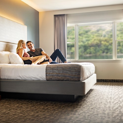 A couple sits on a bed in a hotel room, enjoying each other's company with a window view in the background.