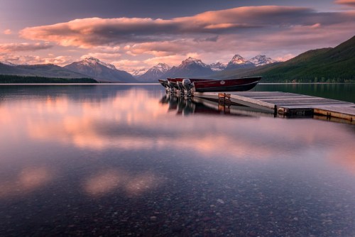 Serene lake at sunset with boats docked, surrounded by mountains and colorful clouds reflecting on the water.