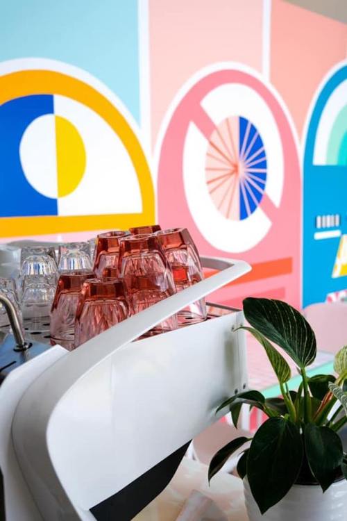 A coffee bar with stacked glass cups and a green plant, set against a colorful, abstract mural.