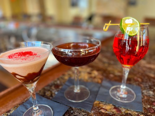 Three colorful cocktails on a bar counter: a chocolate martini, a dark drink, and a vibrant red drink with a lime garnish.