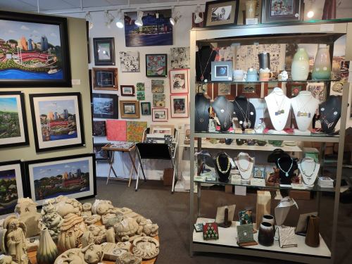 Art gallery display featuring framed artworks, sculptures, and a variety of jewelry on shelves and walls.