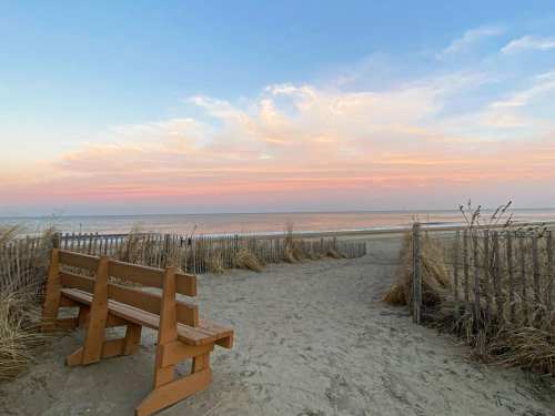 A sandy path leads to a beach at sunset, with a wooden bench and tall grass beside a wooden fence.