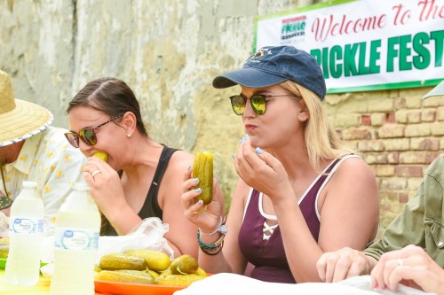 Two women enjoy pickles at a festival, sitting at a table with bottles of water and a "Pickle Fest" banner in the background.