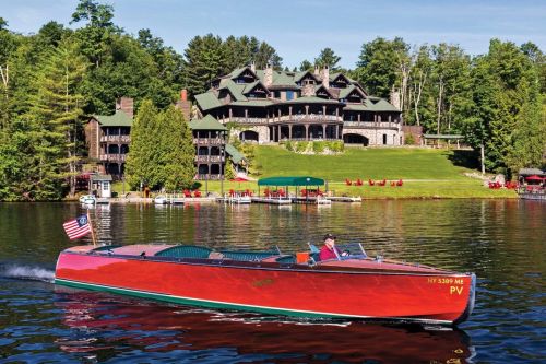 A vintage boat with an American flag glides on a lake near a large, elegant mansion surrounded by trees.