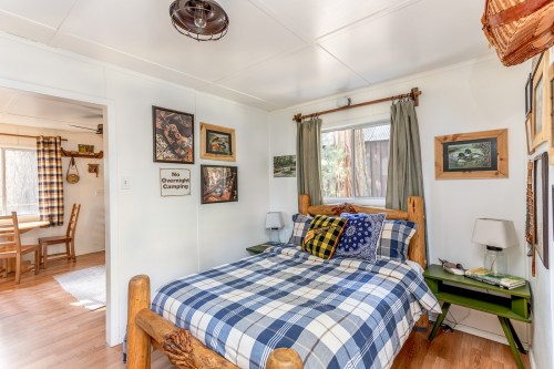 Cozy bedroom with a wooden bed, plaid bedding, and rustic decor, featuring artwork and a small green nightstand.