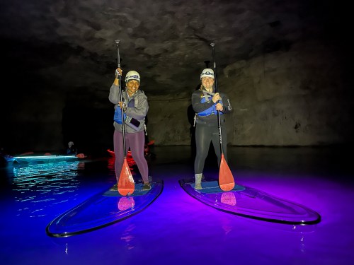 Two women paddleboarding in a dark cave, illuminated by colorful lights reflecting on the water.