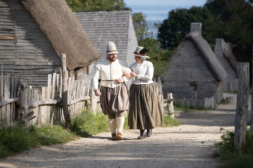 A man and woman in historical attire walk together along a dirt path in a village with thatched-roof houses.