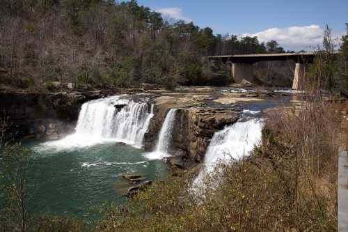 A scenic view of a waterfall cascading into a turquoise pool, with a bridge in the background and trees lining the area.